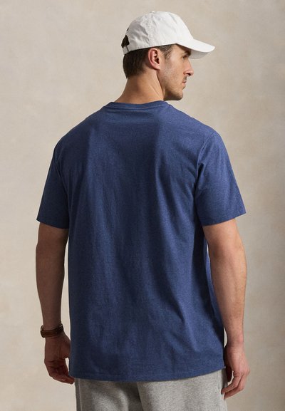 Άνδρας που φοράει απλό μπλε t-shirt, γκρι παντελόνι, λευκό καπέλο μπέιζμπολ και καφέ ρολόι χειρός, γυρισμένος με την πλάτη σε ουδέτερο φόντο.