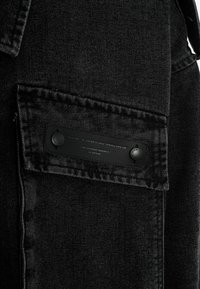 Chaqueta de mezcla de algodón negra con un bolsillo que tiene una etiqueta texturizada y dos botones negros. La tela tiene un acabado liso y ligeramente desgastado.