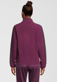 Chaqueta de forro polar púrpura con acentos de tela gris en las mangas; cuenta con un cuello alto y puños elásticos; textura suave y diseño minimalista.
