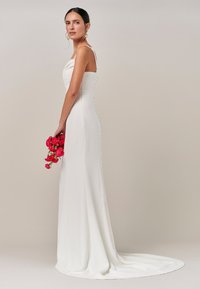 Bela spodnja obleka iz satena z naramnicami iz tankih trakov in s silhueto do tal, z vlečko in gumbnimi detajli ob strani. Drži rožnate rože.