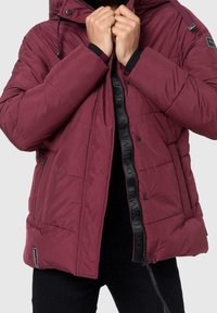 Persoon die een bordeauxrood gewatteerd jack met rits en drukknoopjes draagt en aanpast, met een zwarte binnenvoering en zijzakken.
