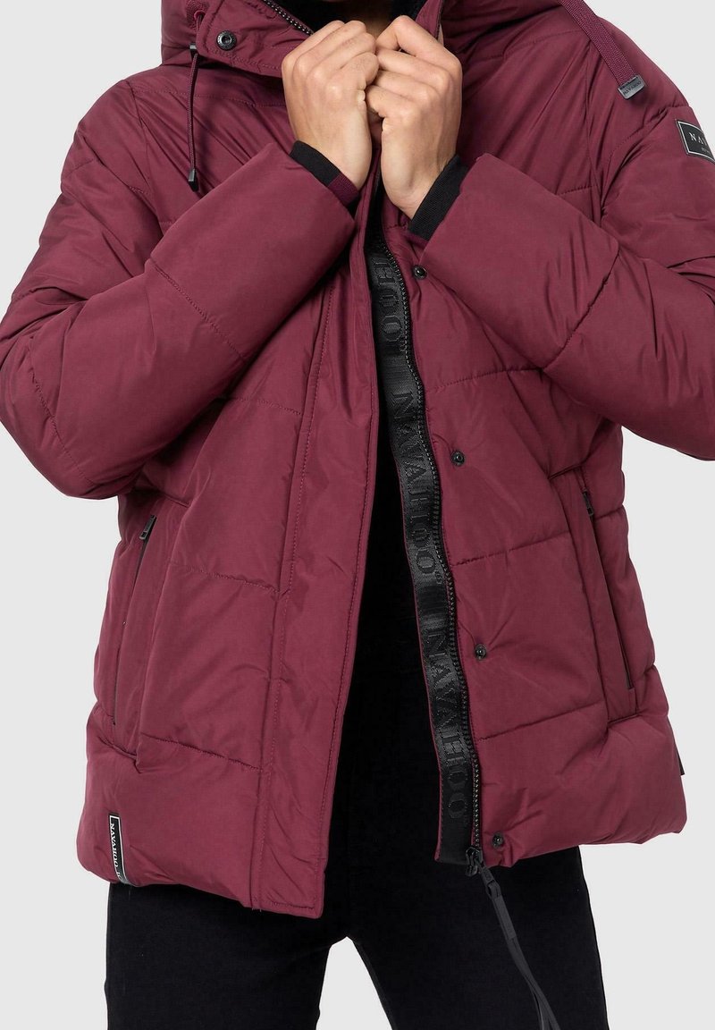 Persoon die een bordeauxrood gewatteerd jack met rits en drukknoopjes draagt en aanpast, met een zwarte binnenvoering en zijzakken.