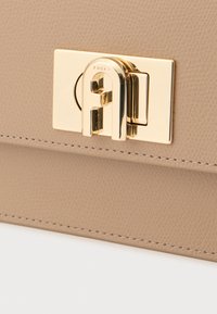 Μπεζ δερμάτινη τσάντα χειρός με χρυσό μεταλλικό κούμπωμα που φέρει το όνομα της μάρκας "Furla". Η υφή είναι ελαφρώς σιαγωνική, με αντιθετικές ραφές.