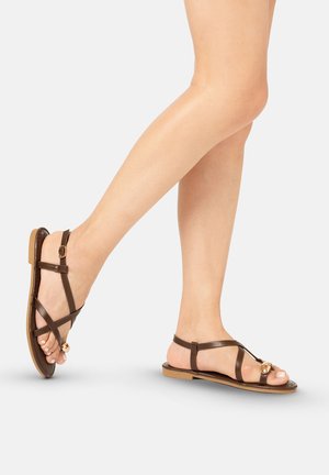 Piernas con sandalias planas marrones con tiras, hebilla en el tobillo y cuenta decorativa dorada en la correa del dedo, posando contra un fondo liso y claro.