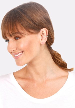 Silberner Ohrstecker am rechten Ohr, mit geschwungener Form und strukturierten Details. Das Model hat kastanienbraunes Haar und trägt ein weißes Oberteil.