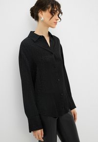 Camicia nera con bottoni, caratterizzata da un sottile motivo leopardato, maniche lunghe e colletto classico. Realizzata in materiale leggero e morbido.