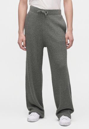 Personne portant un pantalon en maille gris ample avec une taille coulissante et des baskets blanches, debout devant un fond clair uni.
