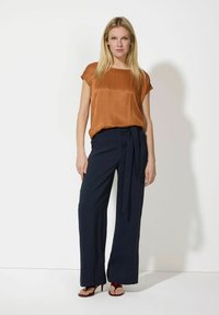 Brązowa satynowa bluzka z krótkimi rękawami w połączeniu z granatowymi spodniami o szerokich nogawkach z wiązanym paskiem. Modelka stoi na neutralnym tle.