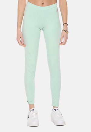 Schmal geschnittene hellgrüne Leggings, getragen von einer Person mit weißen Sneakers, einem Bauchnabelpiercing und Armbändern am rechten Handgelenk.