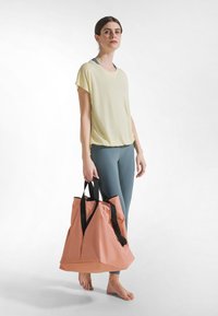 Vaaleankeltainen lyhythihainen toppi, jossa rento istuvuus, yhdistettynä petroolin sävyisiin leggingsseihin. Mallilla on suuri vaaleanpunainen olkalaukku mustilla hihnoilla.