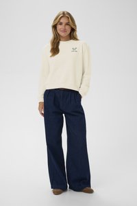 Felpa crema con logo del club di racchetta verde, abbinata a jeans navy a gamba larga e calzature marroni. Texture liscia, design casual.