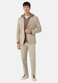 Béžový přizpůsobený blejzr přes hnědou mikinu s pružným zipem, spojený s světle béžovými kalhotami a krémovými teniskami, s uvolněným střihem a minimalistickým designem.