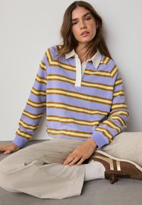 Giovane donna con lunghi capelli castani seduta con le gambe incrociate, indossa una maglietta a maniche lunghe a righe lavanda, senape e bianco e pantaloni beige con scarpe marroni.