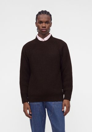 Jeune homme aux cheveux tressés portant un pull en maille marron foncé par-dessus une chemise rose clair à col et un jean bleu, debout devant un fond uni.