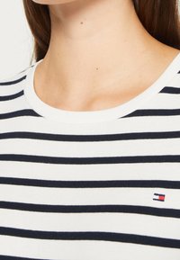 Chemise blanche à rayures horizontales bleu marine, avec un col rond et un petit logo brodé en rouge et bleu sur le bas à gauche.