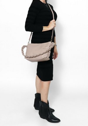 Borsa in pelle morbida di colore rosa con manico intrecciato, dotata di una base piatta. Indossata con una tracolla regolabile, mostrata insieme a stivali alla caviglia neri con frange.