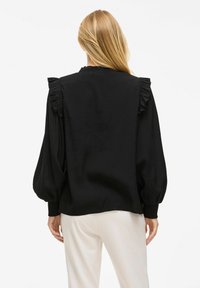 Blouse noire avec des accents froncés sur les épaules, des manches bouffantes longues et une texture douce et lisse ; associée à un pantalon de couleur claire.