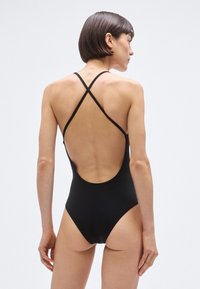 Fato de banho preto de uma peça com costas baixas e alças cruzadas. Tecido liso, design minimalista e aberturas nas pernas com corte alto.