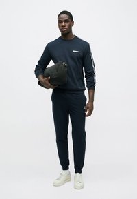 Tmavomodrá mikina a zodpovedajúce tepláky s bielymi pruhmi, doplnené bielymi teniskami. Drží čiernu tašku. Jednoduchý, neformálny dizajn.