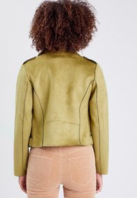 Veste en cuir suédé vert olive avec des coutures noires. Présente des épaulettes et une silhouette ajustée. Texture lisse, détails minimaux.