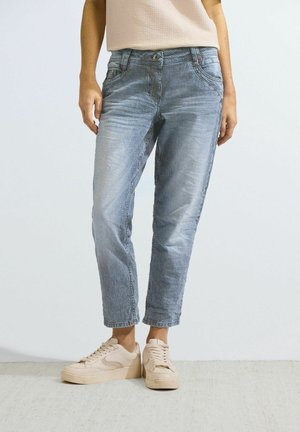 Personne portant un pantalon bleu clair et blanc à fines rayures jusqu'à la cheville avec des baskets beiges, debout sur un fond blanc uni.