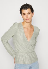 Top envolvente verde claro com padrão texturizado, decote em V profundo, mangas longas com ombros bufantes e faixa elástica na cintura. Combinado com jeans azul.