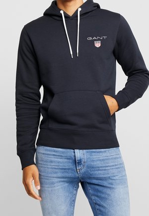 Mand iført en navyblå GANT-hoodie med hvide snore og logo, kombineret med lyseblå jeans, med hånden i hoodie-lommen.