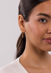 Goldene Creolen mit einem vertikalen Stab-Anhänger, aus poliertem Metall gefertigt, die an einer kleinen Creole hängen und am linken Ohr getragen werden.