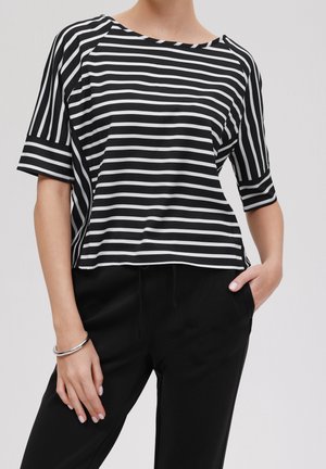 Persona con camiseta de manga corta a rayas negras y blancas y pantalones negros, con una mano en el bolsillo y pulsera de plata en la muñeca.