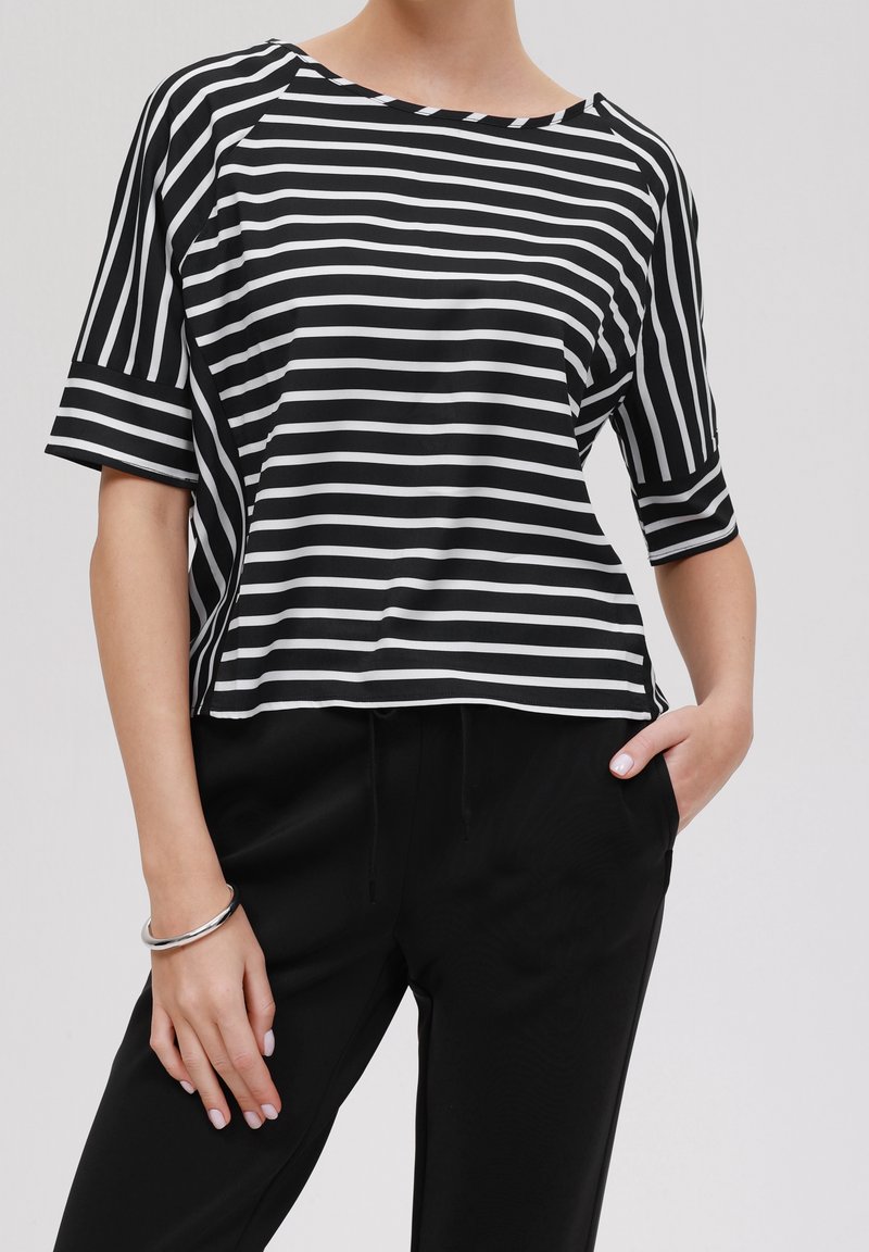 Personne portant un haut rayé noir et blanc à manches courtes et un pantalon noir, avec une main dans la poche et un bracelet argenté au poignet.