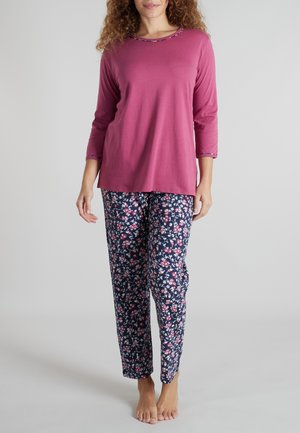 Frau trägt ein langärmeliges, rosarotes Oberteil und marineblaue Pyjamahosen mit floralem Muster, steht barfuß auf einem hellgrauen Boden.