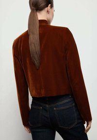 Giacca in velluto marrone cropped con maniche lunghe, texture morbida e collo alto. Indossata sopra jeans in denim scuro, che mettono in risalto una silhouette elegante.