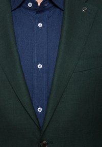 Ciemnozielona teksturowana marynarka nałożona na ciemnoniebieską koszulę dżinsową z kołnierzykiem zapinanym na guziki, z jasnoniebieskimi guzikami i przeszyciami.