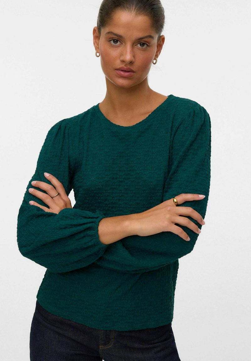 Teal, texturerad blus med långa ärmar och puffärmar, rund halsringning, stylad med mörka jeans. Subtilt mönster genom tyget.