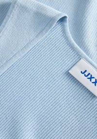 Ανοιχτό μπλε πλεκτό ύφασμα με ριμπ υφή, τετράγωνο ντεκολτέ και λευκή υφασμάτινη ετικέτα με μπλε κείμενο, που φαίνεται μερικώς.