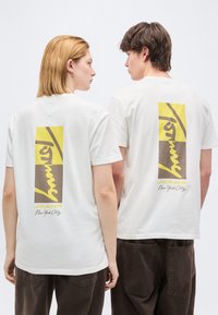 Δύο άτομα φοράνε λευκά γραφικά μπλουζάκια με κίτρινες και καφέ ορθογώνιες ετικέτες και το κείμενο «JEANS New York City» στην πλάτη τους.