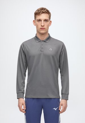 Jeune homme portant un polo gris à manches longues et un pantalon de sport bleu avec des logos, debout et faisant face à un fond uni.