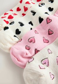 Trei perechi de șosete: una albă cu inimioare roșii, una crem cu inimioare negre și una roz cu inimioare roz mai deschis, toate din material tricotat moale.