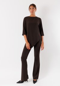 Maglia tunica marrone con maniche tre quarti, orlo asimmetrico, abbinata a pantaloni marroni aderenti e tacchi neri a punta. Tessuto dalla consistenza liscia.