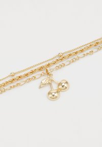 Collier doré à plusieurs couches avec un pendentif cerise, deux charms sphériques et un détail de feuille, chaînes entrelacées texturées.