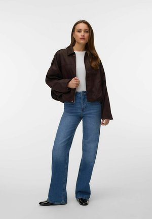 Žena stojaca v čiernej zapínacej bunde cez bielu košeľu, modrých džínsach s vysokým pásom a širokými nohavicami a čiernych topánkach na bielom pozadí.