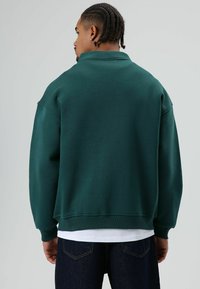 Groene oversized sweatshirt met een hoge kraag, geribbelde manchetten en zoom. Het materiaal lijkt zacht en gestructureerd, gedragen over een wit T-shirt.