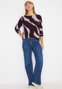 Sweter dzianinowy w kolorze burgundowym i beżowym z wzorem, z rękawami 3/4, zestawiony z niebieskimi spodniami o szerokich nogawkach i beżowymi trampkami.