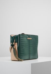 Borsa a mano verde con effetto coccodrillo, con base beige, dotata di zip dorata e dettaglio con frangia. Forma rettangolare con design strutturato.