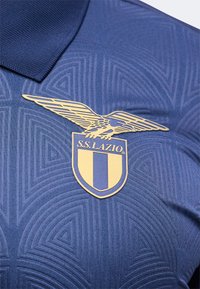 Μπλε αθλητική φανέλα με χρυσό έμβλημα αετού και κείμενο "S.S. LAZIO" στο στήθος, με γεωμετρικά υφασμάτινα μοτίβα.