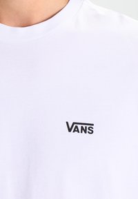 Valkoinen puuvillainen T-paita, jossa vasemmassa rintakehässä musta "VANS"-logo; pyöreä kaula-aukko ja pehmeä materiaali.
