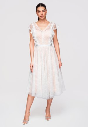 Donna che indossa un abito midi bianco, trasparente, a pois, con maniche a cappuccio arricciate e scollo a V, abbinato a tacchi aperti.