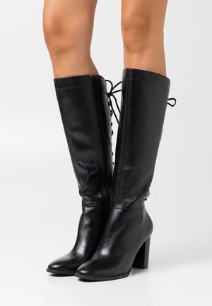 Bottes hautes jusqu'aux genoux en cuir noir au toucher lisse, fermeture éclair latérale et détail contrastant à lacets à l'arrière. Talon bloc.
