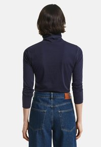 Persoon met kort bruin haar, gekleed in een marineblauwe langmouwen coltrui en hoge blauwe denim jeans, gezien vanuit de rug.