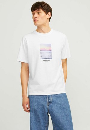 Ung mann som ser til siden, iført en hvit T-skjorte med et pastellfarget havsolnedgangstrykk og blå jeans mot en ensfarget bakgrunn.